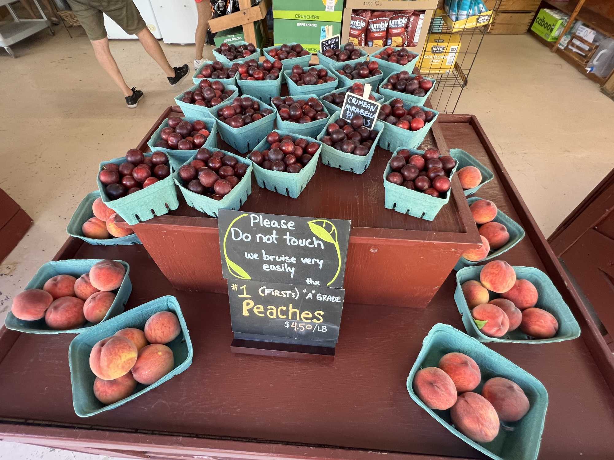 Baskets of fresh-picked peaches and Crimean Mirabelle plums displayed on a farm stand table in the Similkameen Valley, BC, with a handwritten chalkboard sign reading "Number 1 A Grade Peaches $4.50/lb" — the kind of roadside fruit stand found along Highway 3 between Keremeos and Cawston.