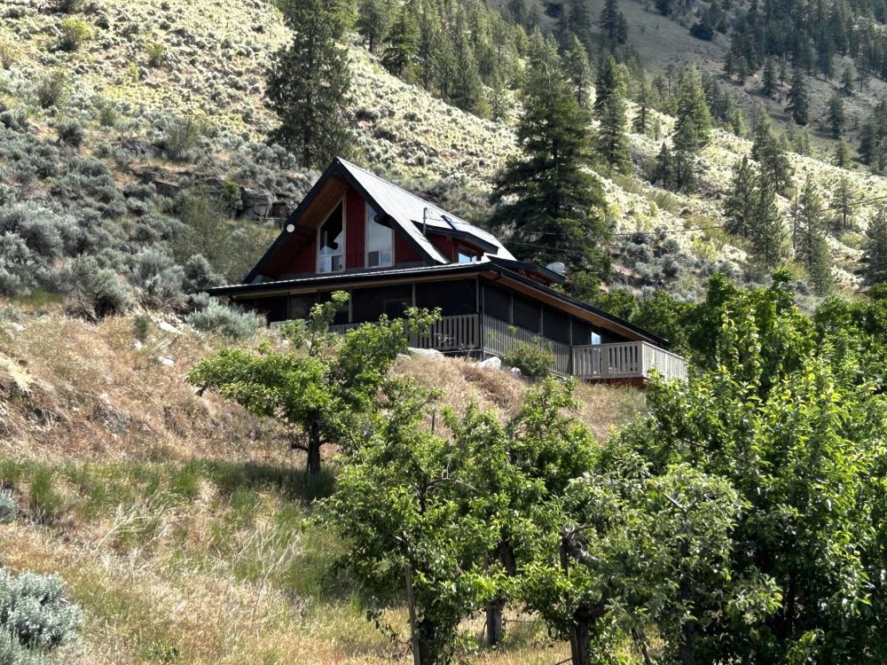Log cabin guesthouse nestled on a hillside among fruit trees and sagebrush, with a wraparound deck and pine-forested slopes behind